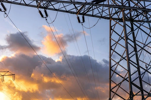 High voltage electrical transmission tower and power lines silhouetted against a cloudy sunset sky.