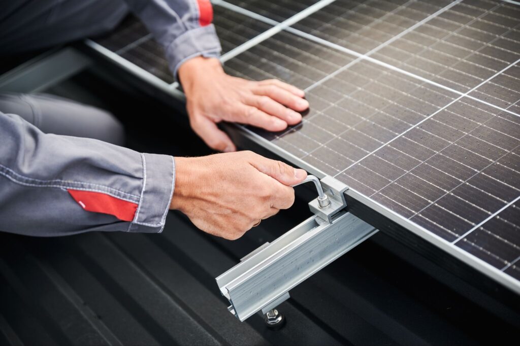 Close-up of a worker's hands using an Allen wrench to tighten a metal clamp, securing a wet solar panel to an aluminum mounting rail on a dark corrugated roof.