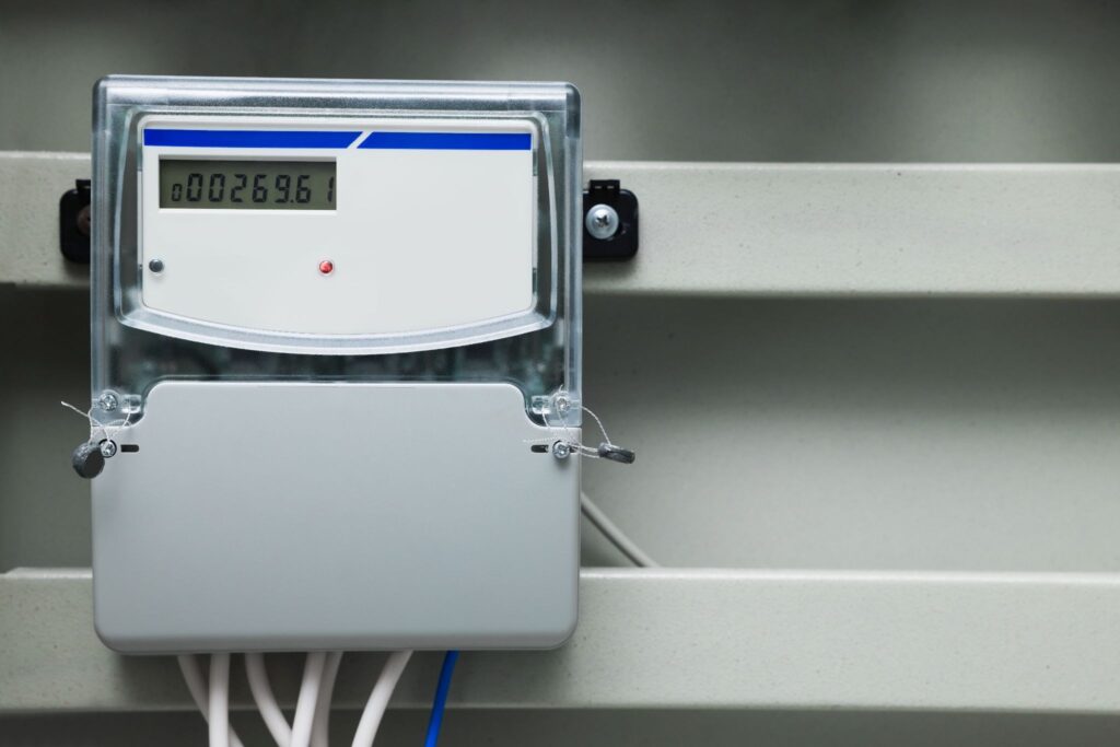 Close-up of a digital electric meter mounted on a grey metal box, featuring a digital display showing the number 00269.61, a small red indicator light, and wires extending from the bottom