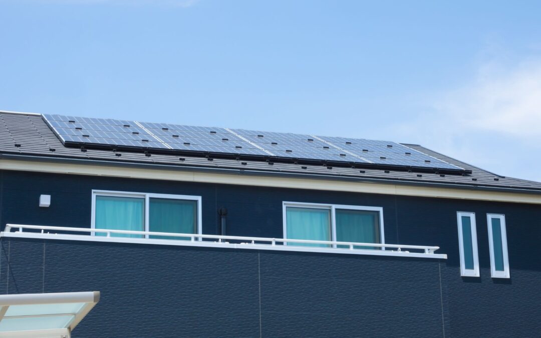 A modern house featuring a large array of solar panels installed across its roof, a white balcony railing, and sliding windows against a clear blue sky.